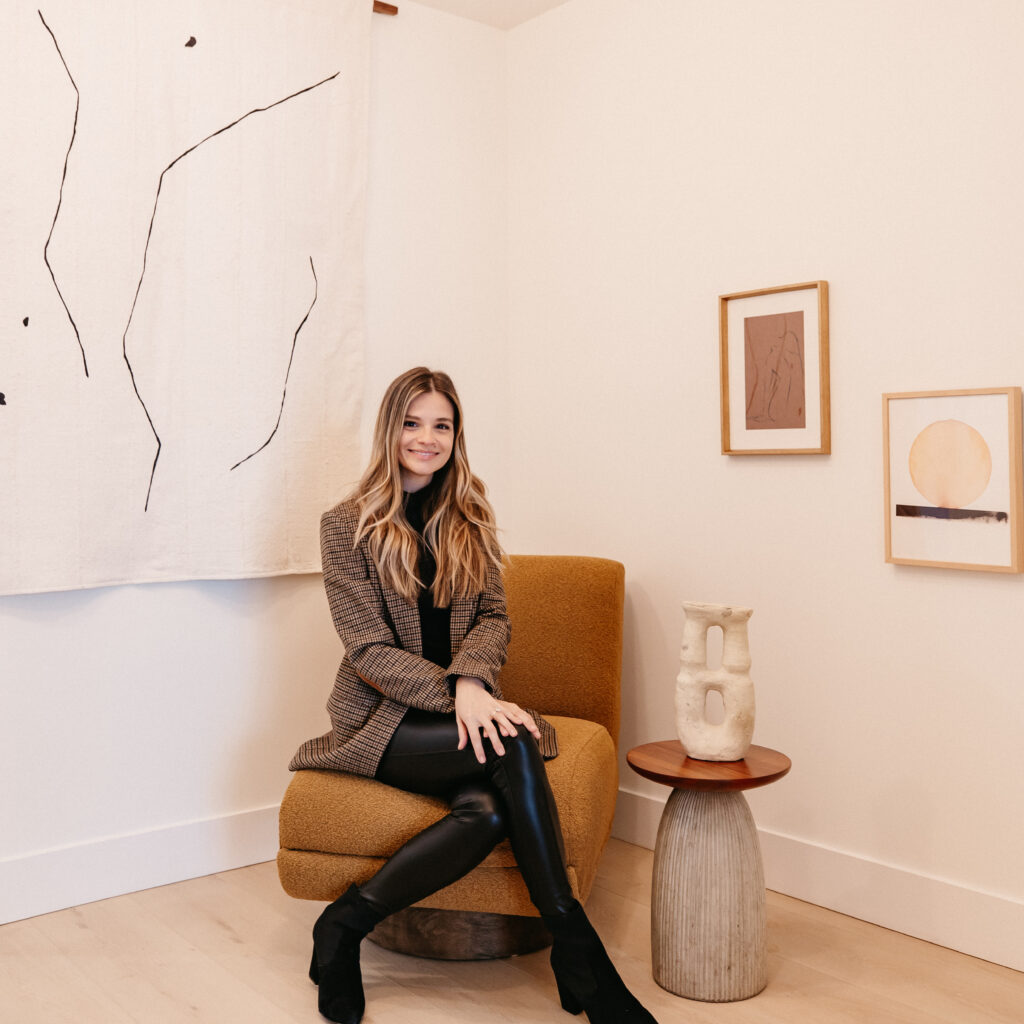 Dr. Vickey seated in a modern wellness clinic lounge, wearing a blazer and leather pants, surrounded by minimalist art and warm neutral decor.