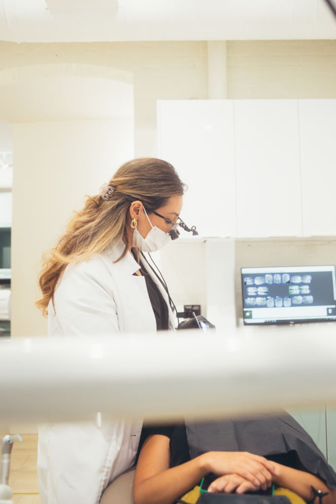 Female dentist providing dental treatment to a patient in a modern dental clinic, wearing magnification loupes and a protective mask